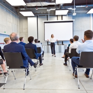 In a large room, people in chairs look at a woman presenting in front of a projection screen.