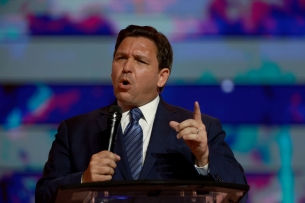 Ron DeSantis, wearing a suit and tie, pointing and talking into a microphone