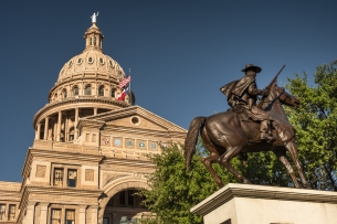 The Texas Capitol building looms behind a statue of a man on a horse with a gun.