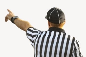 A referee in a black and white striped shirt and a black baseball cap, faces away from the camera as he points toward the distance.