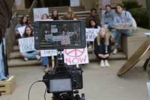 A camera films students with signs on steps