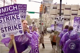 People, most wearing purple ponchos, hold purple and white signs on a sidewalk. The signs say "UMich Grad Workers Strike Strike Strike" and the union's name.